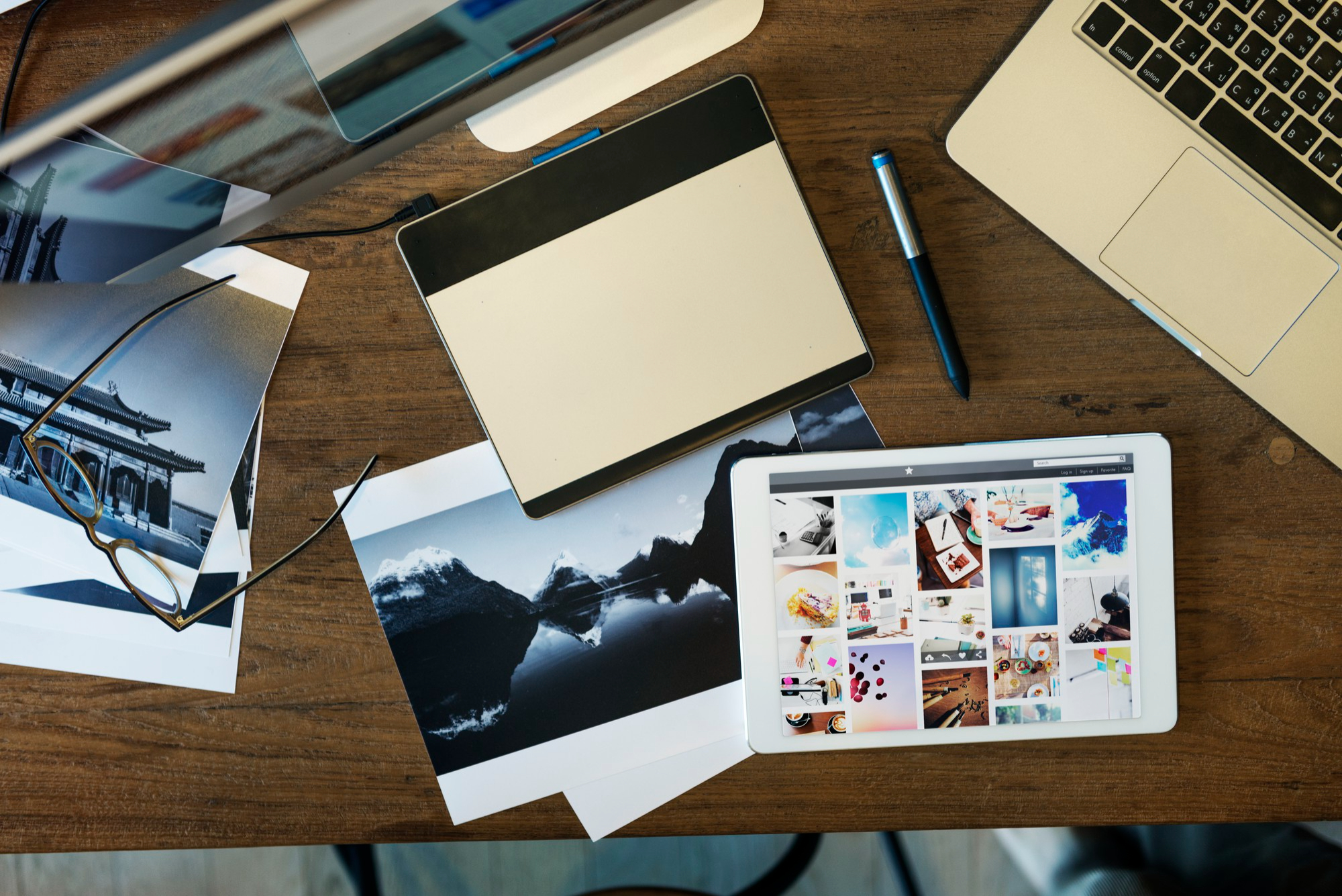 Wooden desk with a digital drawing tablet, a white tablet displaying photos, a laptop keyboard, a pen, printed photographs, and eyeglasses.