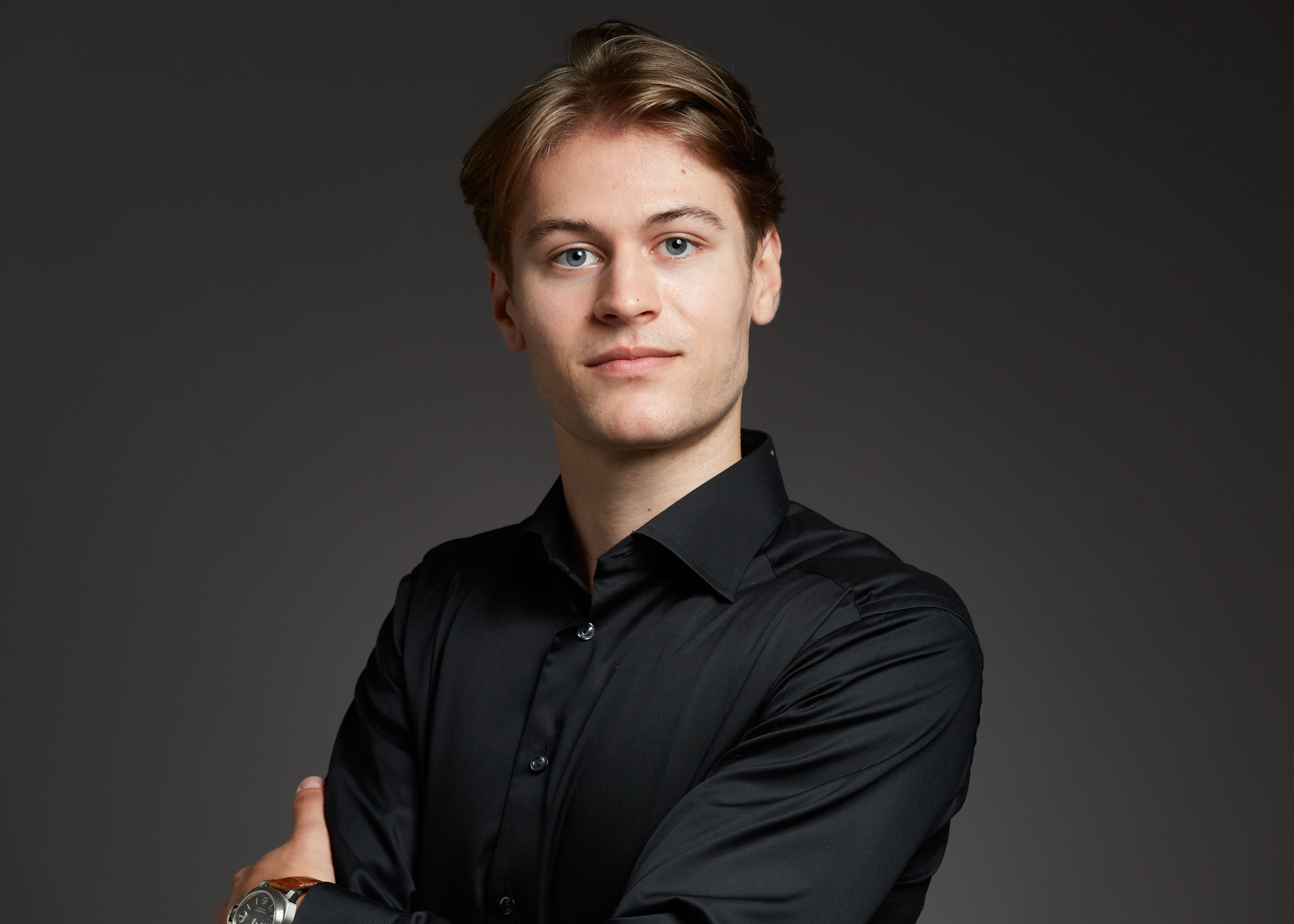 Young man with light brown hair wearing a black button-up shirt and a wristwatch, standing with arms crossed against a dark background.