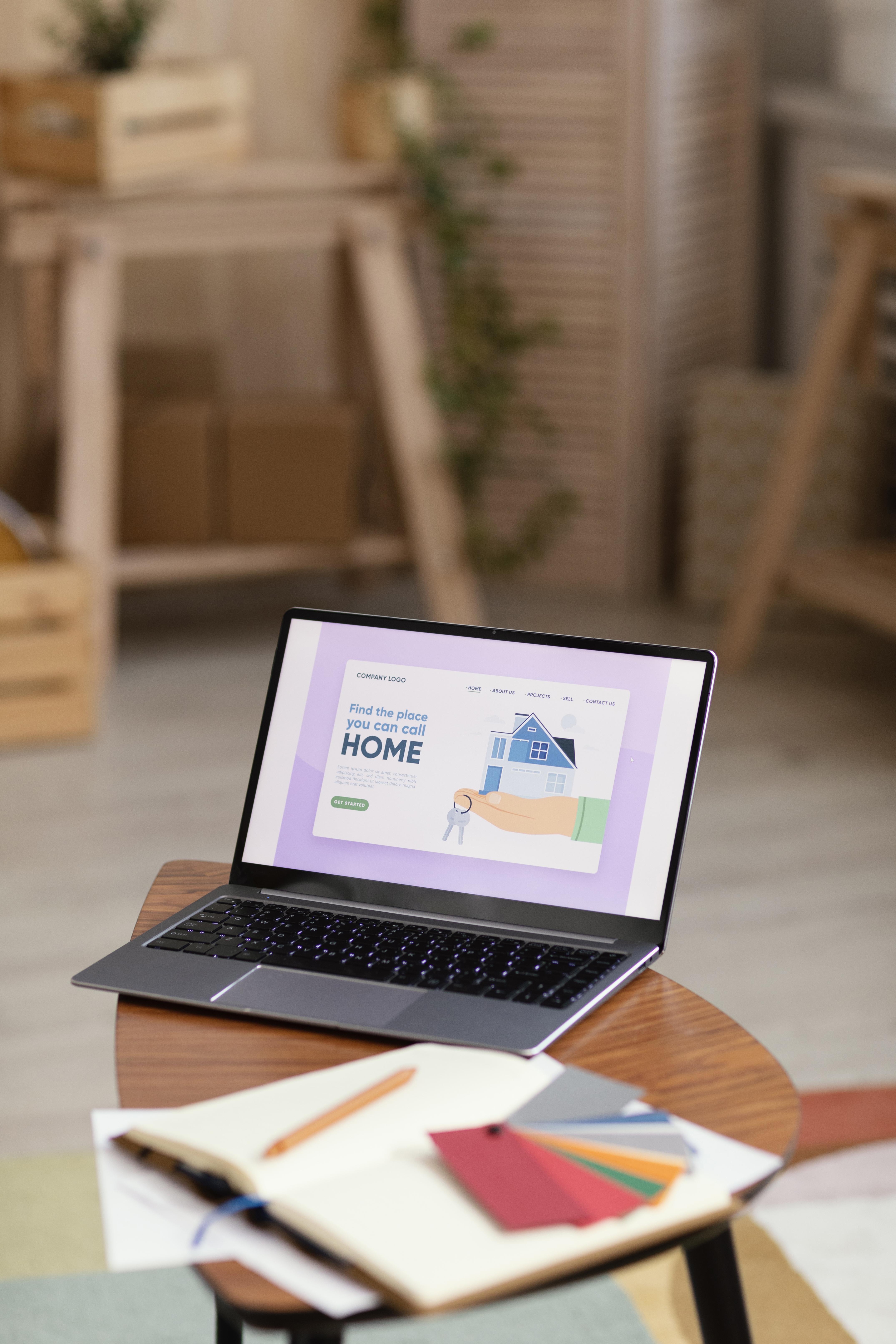 Laptop on a wooden table displaying a website with the text 'Find the place you can call HOME' alongside an illustration of a hand holding a house and keys.