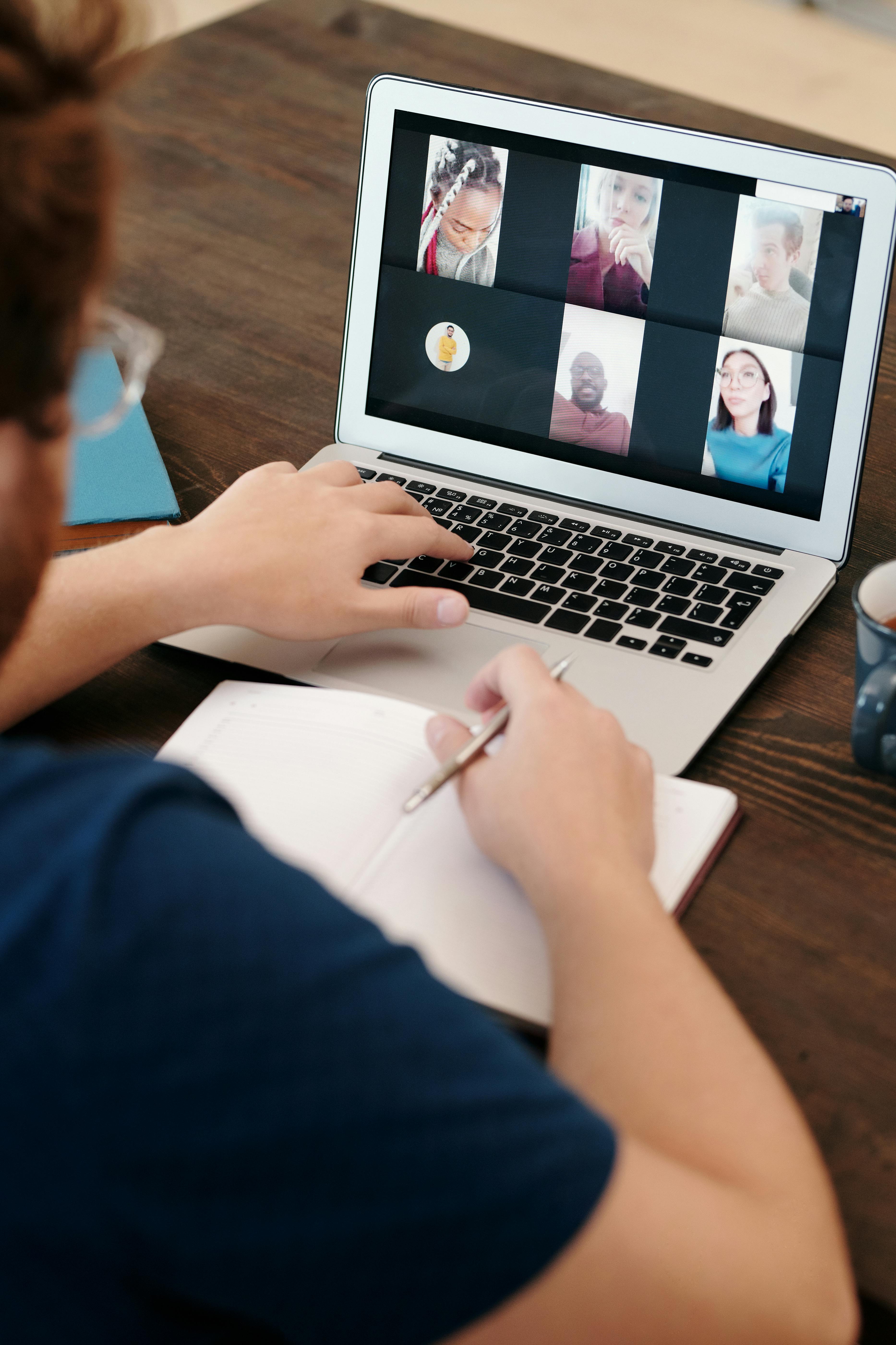 Person attending a video conference on a laptop while taking notes in a notebook at a wooden desk.