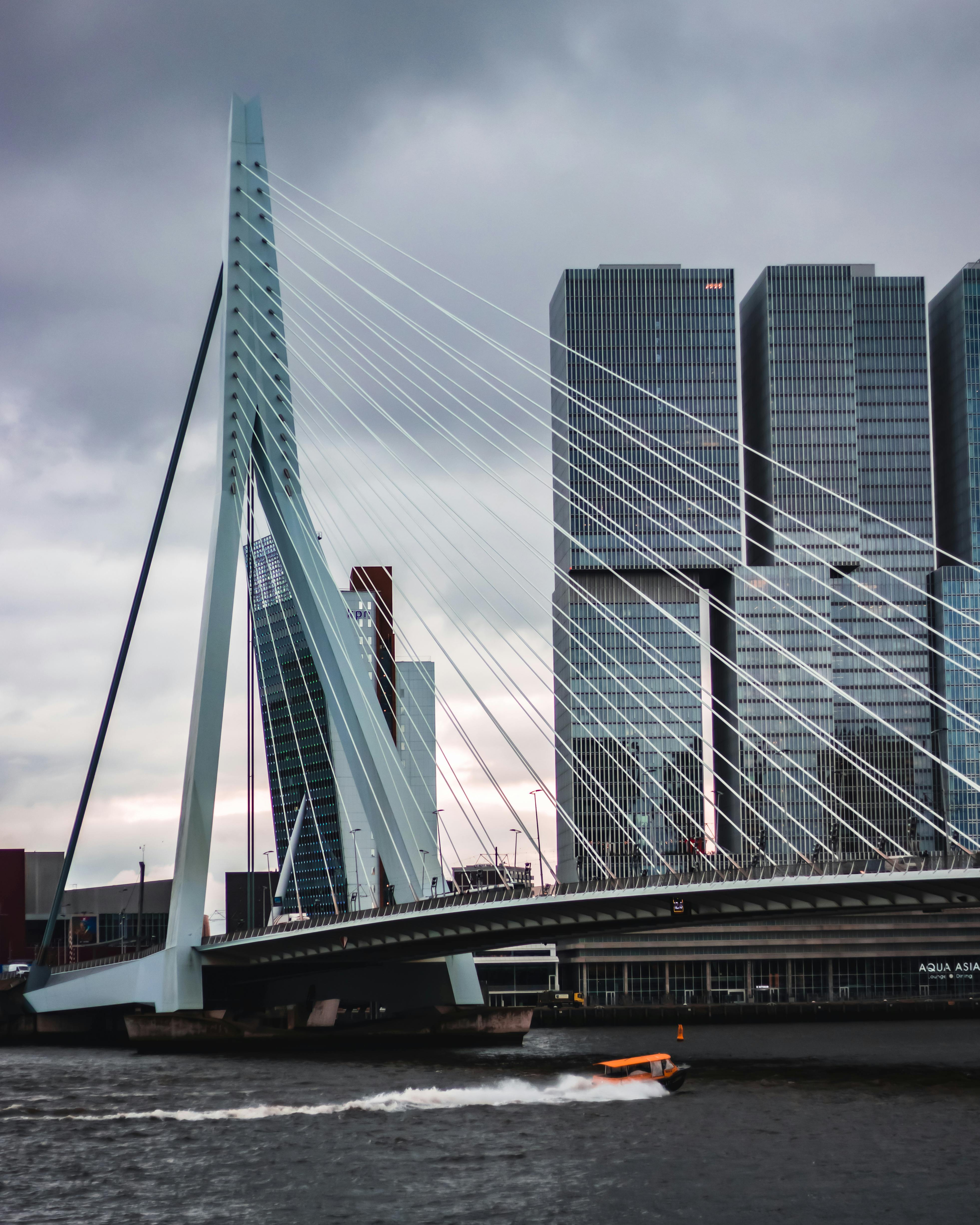 Modern cable-stayed bridge over a river with a small orange boat speeding beneath and tall glass skyscrapers in the background under a cloudy sky.